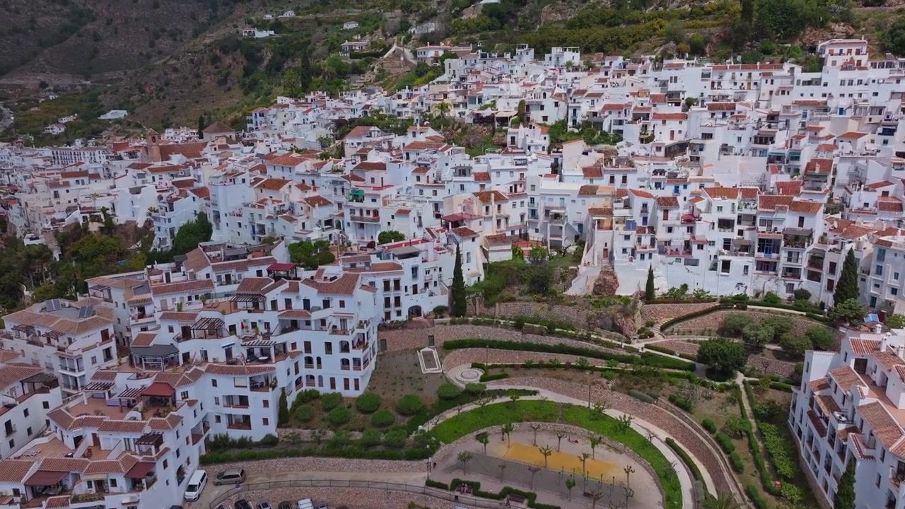 Droneoptagelse af Frigiliana, Andalusien, der viser den centrale plads, hvidkalkede huse på bjergskråningen og dramatisk bjerglandskab i et bredt filmisk oversigtsbillede