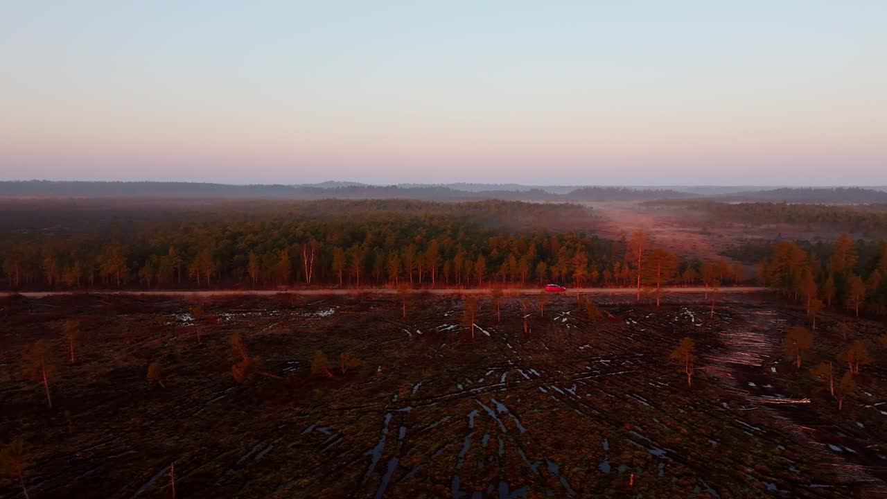 Red car slowly drives in reverse on gravel road by misty Raganu Swamp at sunrise