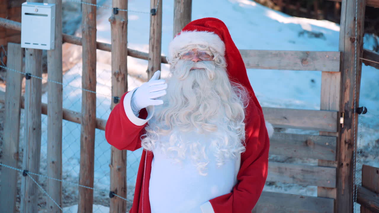 산타가 나무 울타리 옆에 서서 말하고 카메라를 향해 손을 흔들고 있다