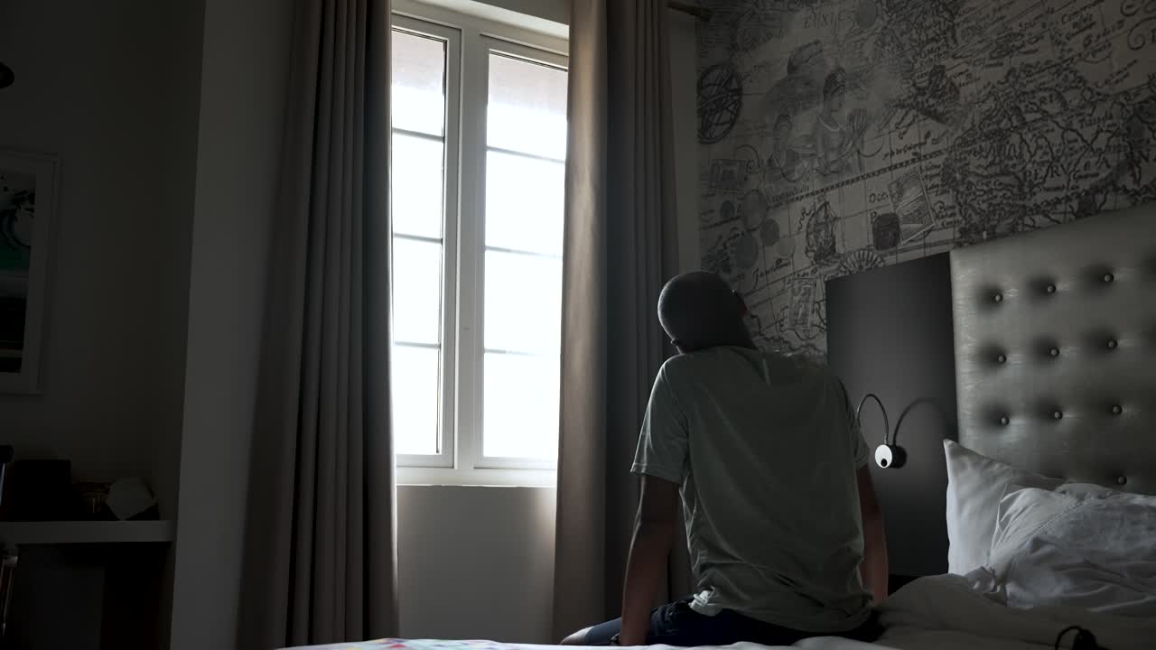 A man sits on a bed in a hotel room, stretching towards the bright window, conveying relaxation and travel mood.