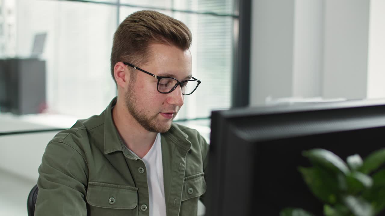 charmanter mann mit brille für das sehen arbeitet am computer, während er am tisch im büro sitzt. hintergrund des fensters