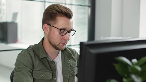 charming man in glasses for vision works at computer while sitting at table in office background of window