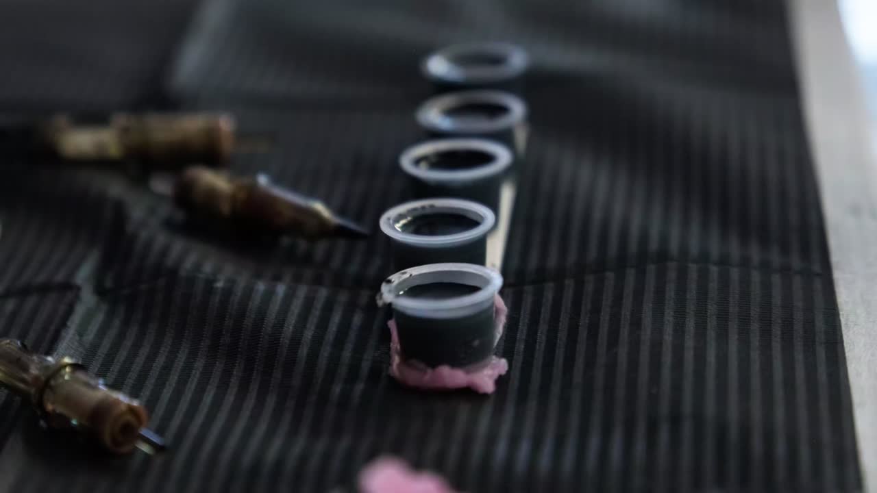 Tattoo artist dips tattoo gun into black ink in cup inside tattoo studio.