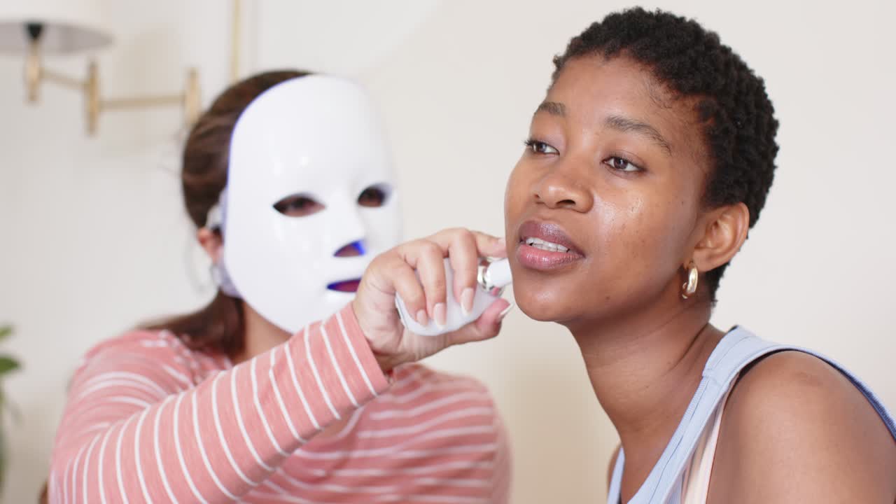 En kvindelig behandler fører et LED-maske-apparat fra håndled til kæbe på en afroamerikansk kvinde til en ansigtsbehandling derhjemme.