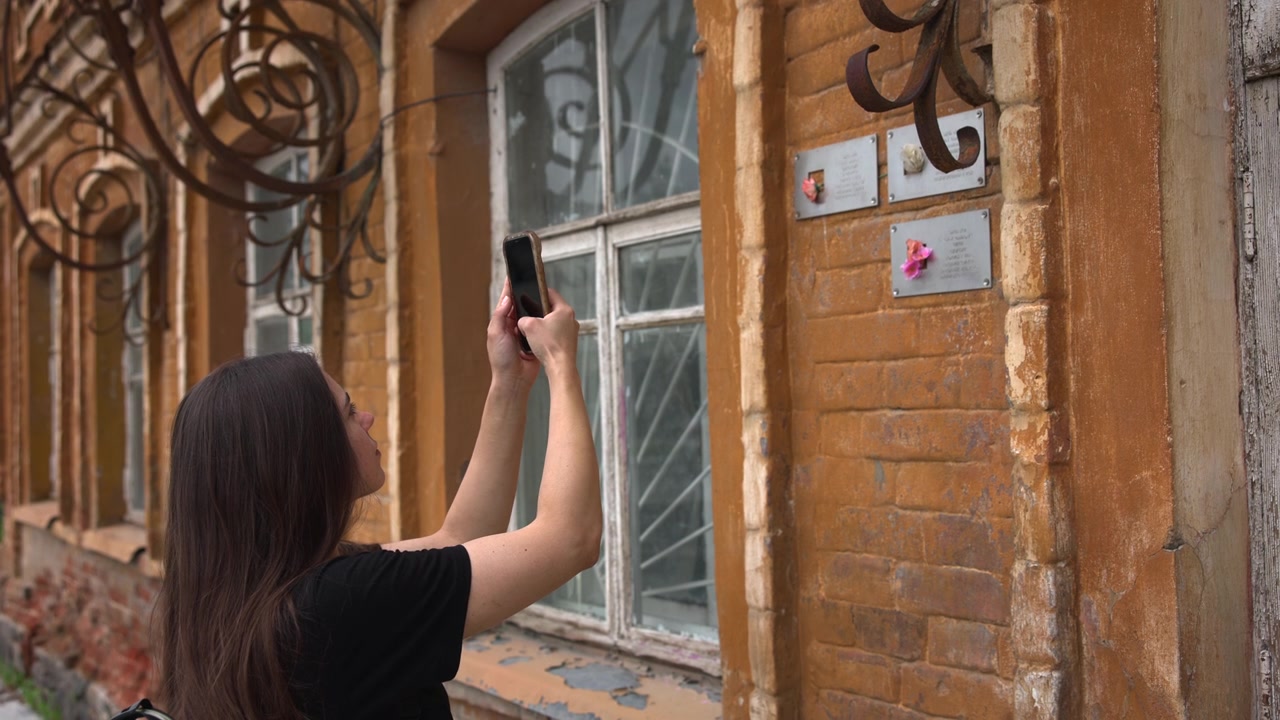 Rear side view shows young woman with long dark hair pausing beside heritage brick wall, looking down at smartphone after photographing aged facade details during quiet urban daylight