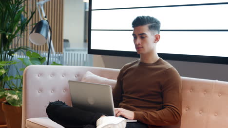 Close-up-of-millennial-businessman-working-on-a-laptop-computer-sitting-on-a-sofa-in-a-casual-office,