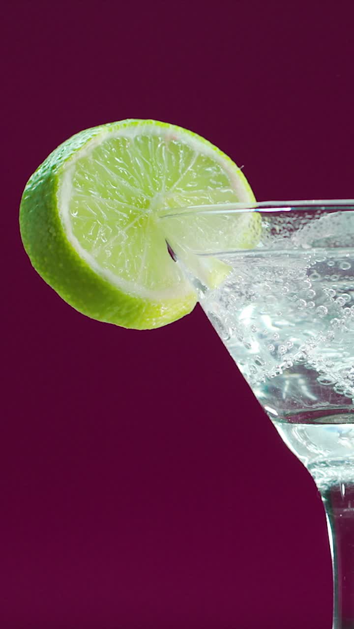 Clear sparkling drink in martini glass with lime slice rotating on bordeaux background