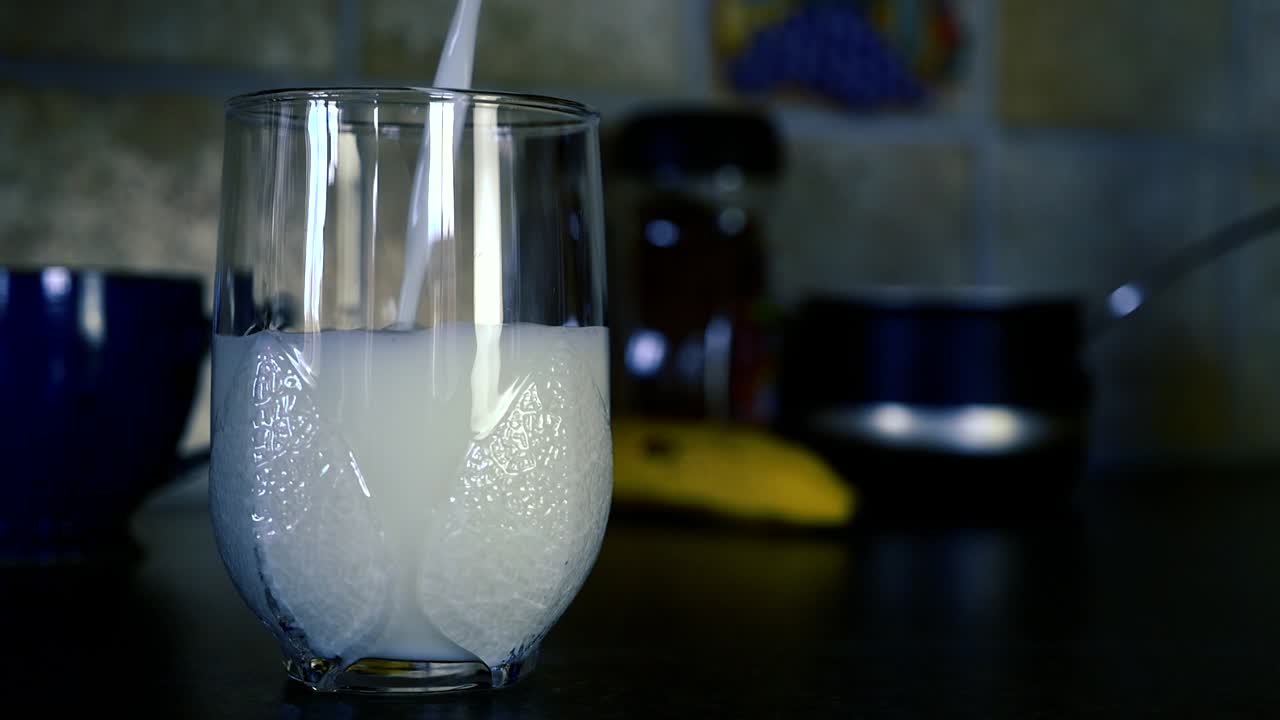 Milk is poured in an empty glass with fruits and kitchen equipment in background