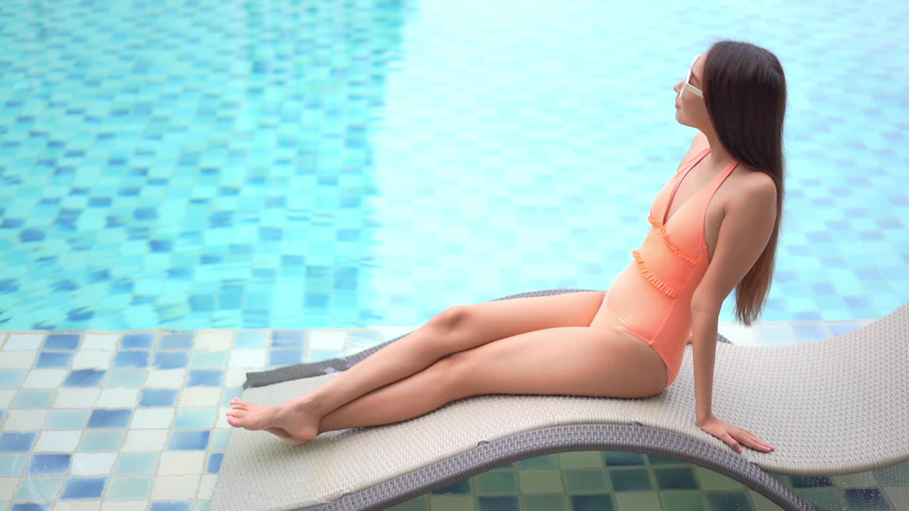 A young pretty woman relaxes on a rattan lounger alongside a resort swimming pool
