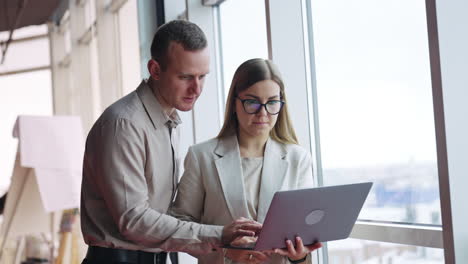 male and female colleagues discuss something on laptop. woman is holding a notebook and points at something. man is using touch pad to search the information.
