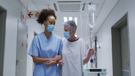 Female-diverse-doctor-and-patient-wearing-face-masks-walking-through-hospital-corridor