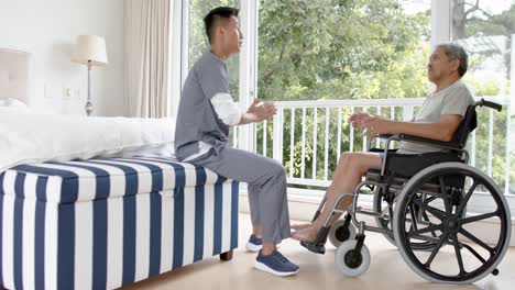 diverse male doctor and senior male patient in wheelchair discussing at home, slow motion