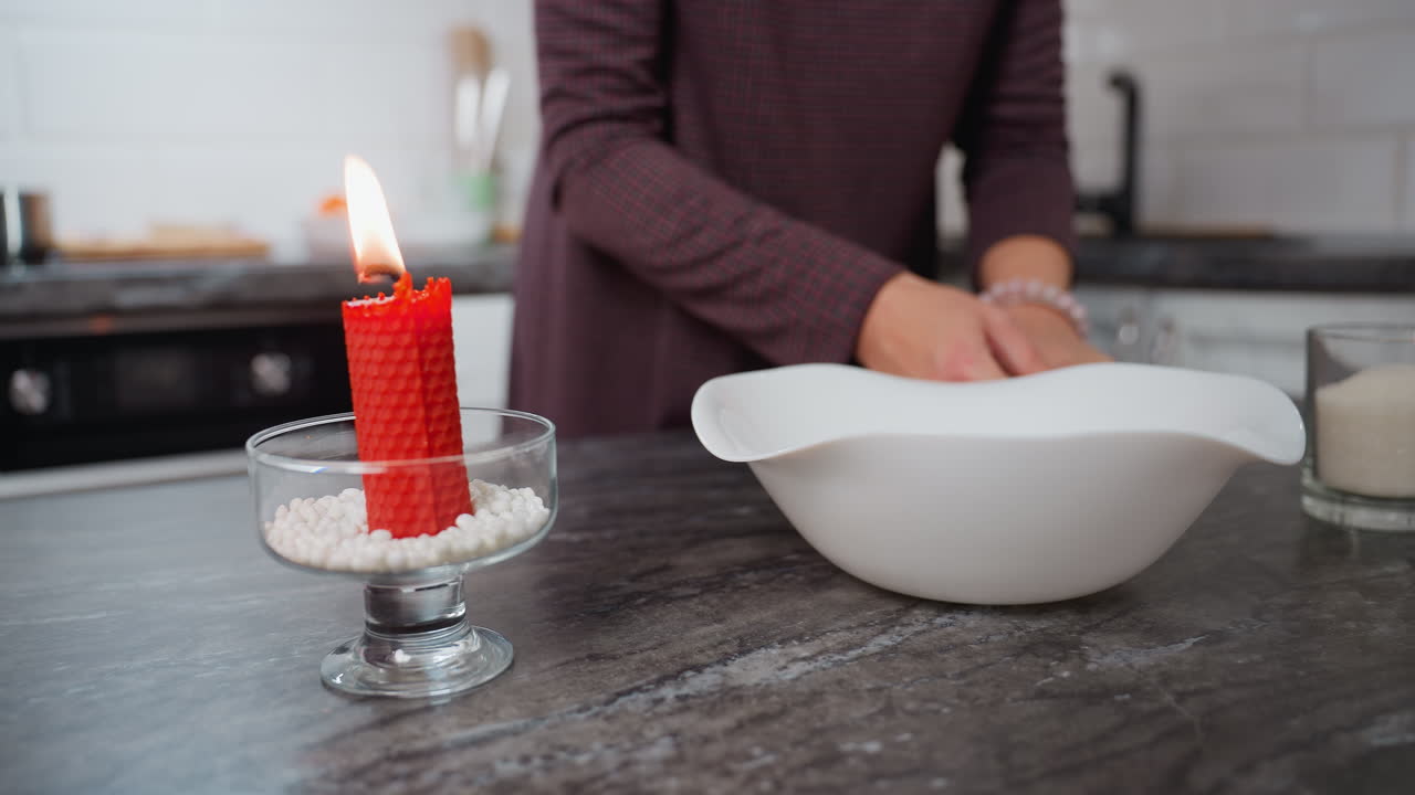 vista parcial de una mujer colocando un cuenco blanco en la mesa de la cocina mientras sostiene huevos en la mano adornados con una pulsera, una vela roja ardiente y una taza de vidrio de azúcar en la mesa