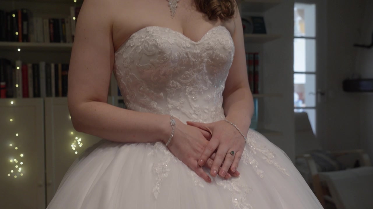 Sposa in abito da sposa bianco in piedi all'interno con le mani giunte, gioielli eleganti visibili.