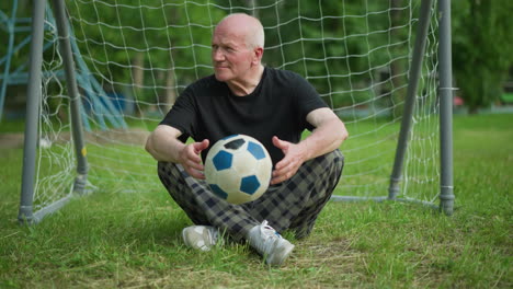 an elderly man sits cross-legged near a goal post, rotating a soccer ball in his hands, with trees in the background