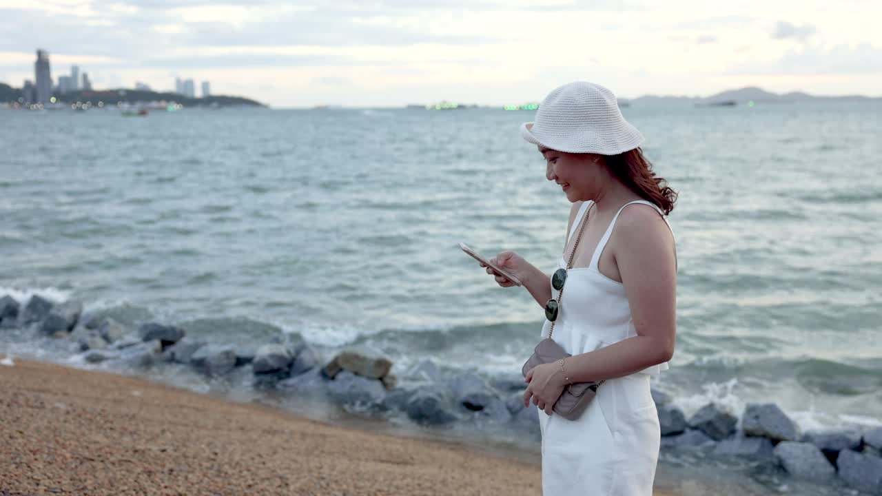 아시아 여성이 해변에 서서 휴대전화로 친구들과 채팅하고 있습니다.