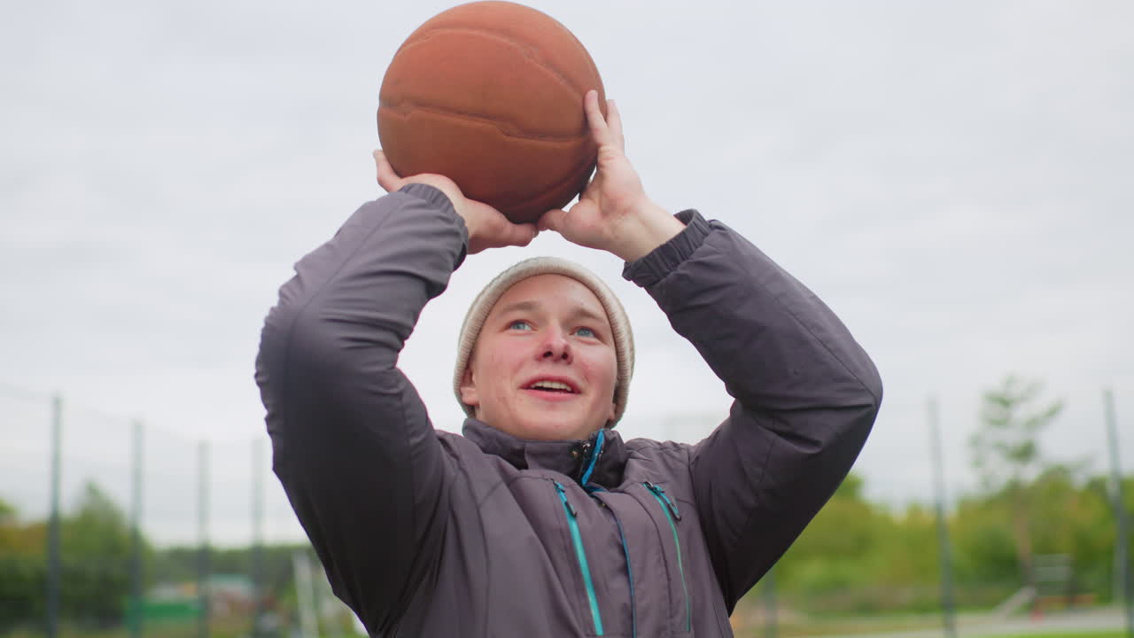 teen shooting basketball on outdoor court, beanieclad player lines up shot under overcast sky, careful aim and release, followthrough captured midair, background shows fenced park and distant