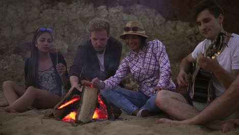 groep jonge en vrolijke mensen die 's avonds bij het vuur op het strand zitten, liedjes zingen en gitaar spelen.