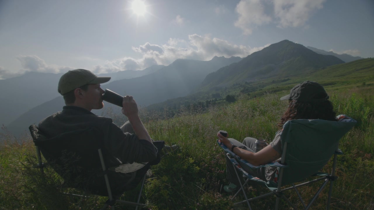Couple sit overlooking mountain scenery on a sunny morning drinking tea from flask in the alps in summer.