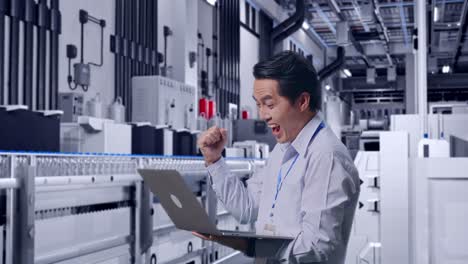 side view of an asian male professional worker standing with his laptop with water production in bottling factory, he raises his fist up with screaming goal after typed on the laptop