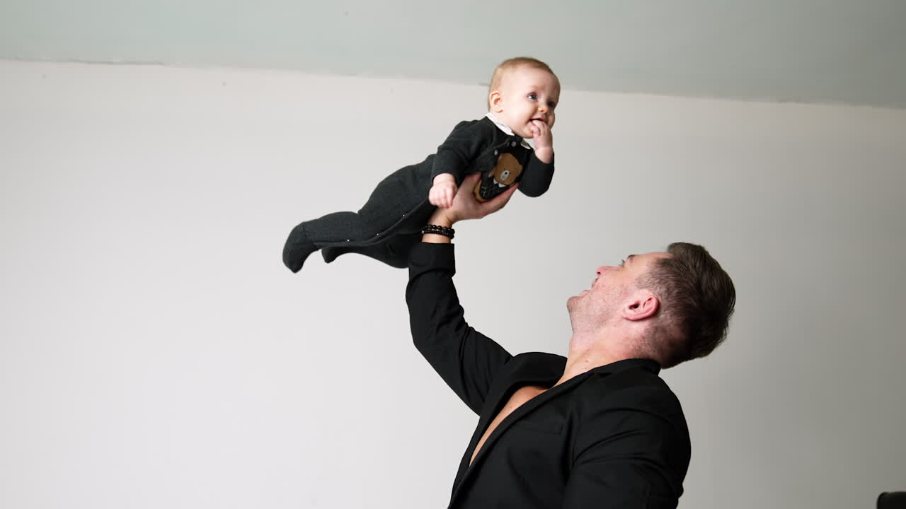 Strong Caucasian man in black jacket raises a baby with one hand. Father is playing with his little son. White backdrop.
