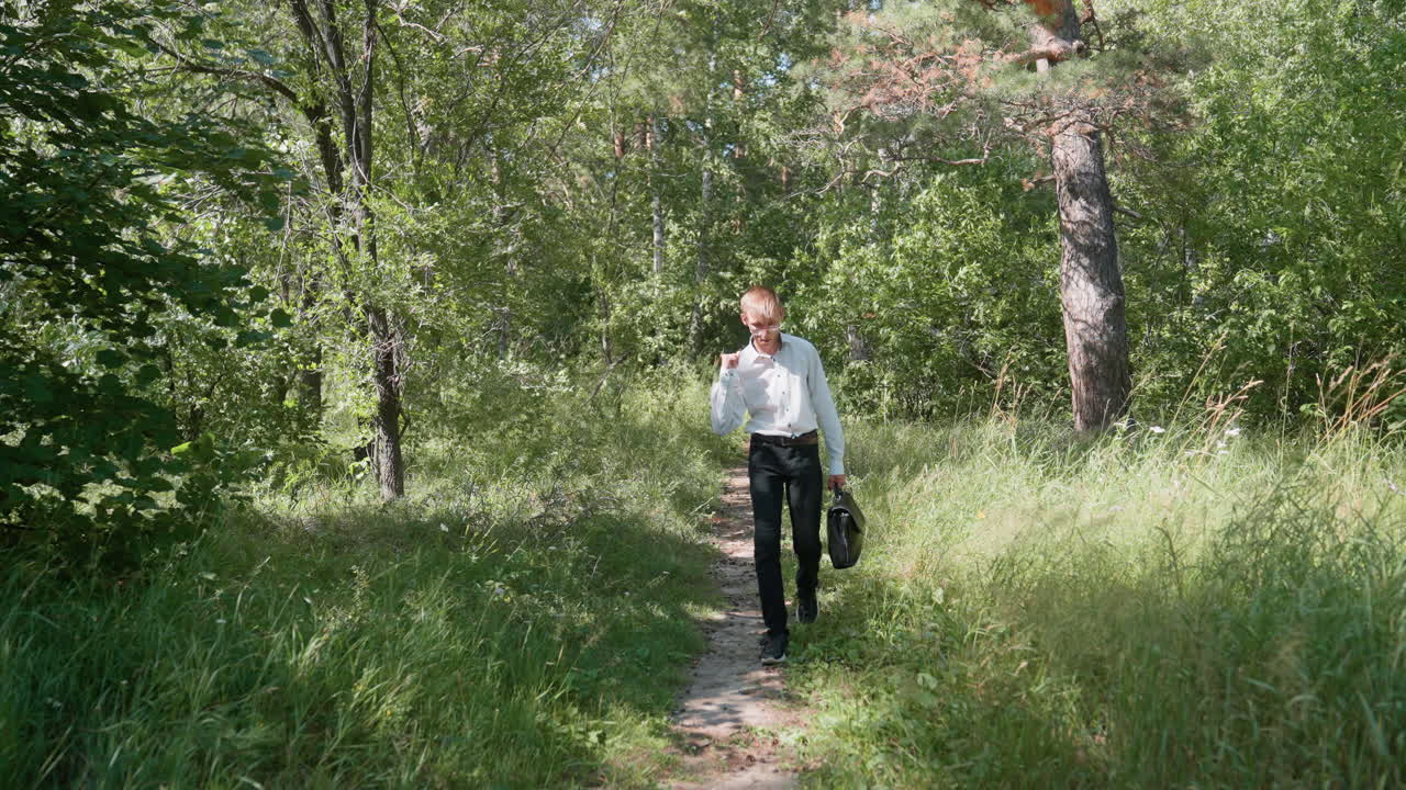 Man in white shirt and black trousers carrying backpack walking slowly along forest path, smiling as he carefully observes plants surrounded by lush greenery and sunlight streaming through tall trees