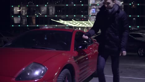 man getting into red sport car on drivers side. city lights at night on the background