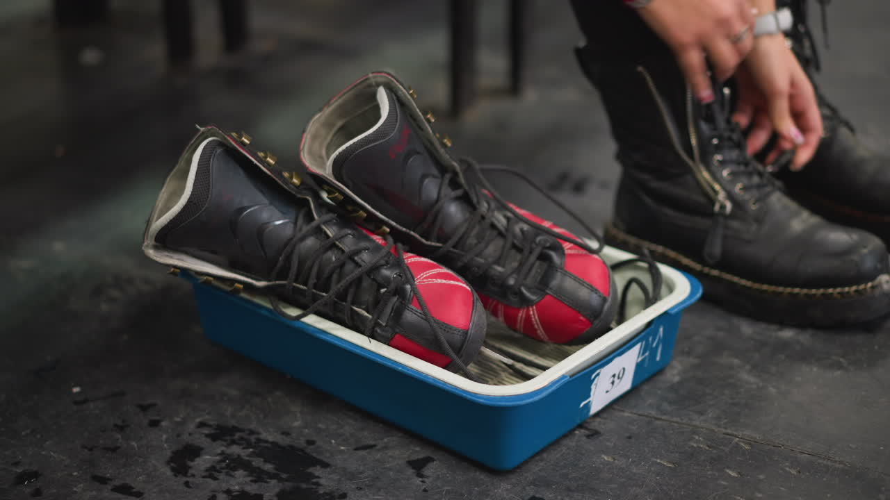 Female placing pair of red and black ice skates in plastic tray on dark floor with hands visible wearing bracelets, changing shoes preparation for skating activity winter sports