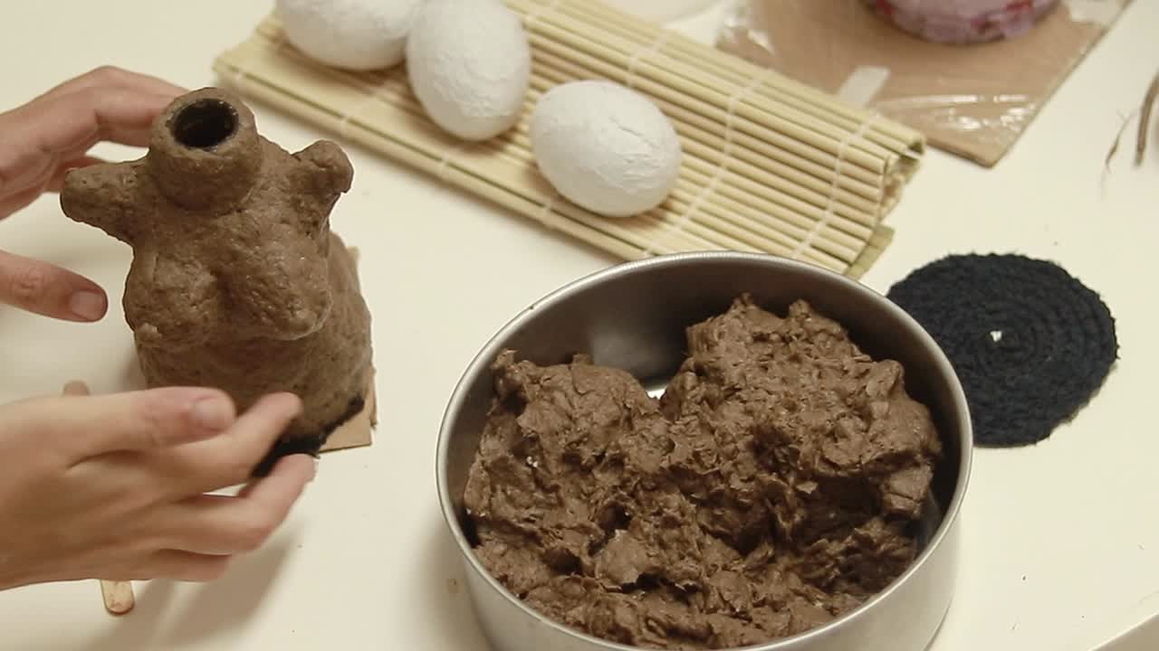 Hands shaping paper air dry clay made of recycled egg carton beside a pottery piece on a crafting table, DIY craft project