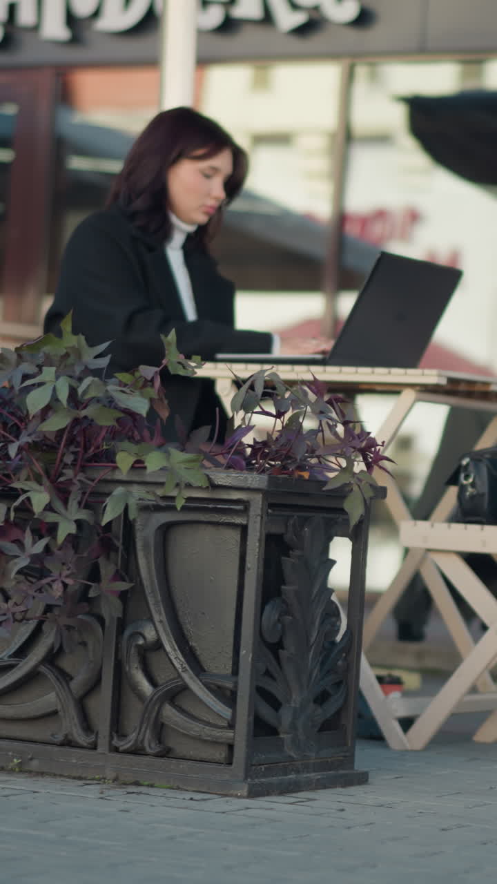 Student with dark hair typing on laptop at outdoor table in cozy mall setting, surrounded by plants and chairs with blurred background featuring a visible inscription on the wall