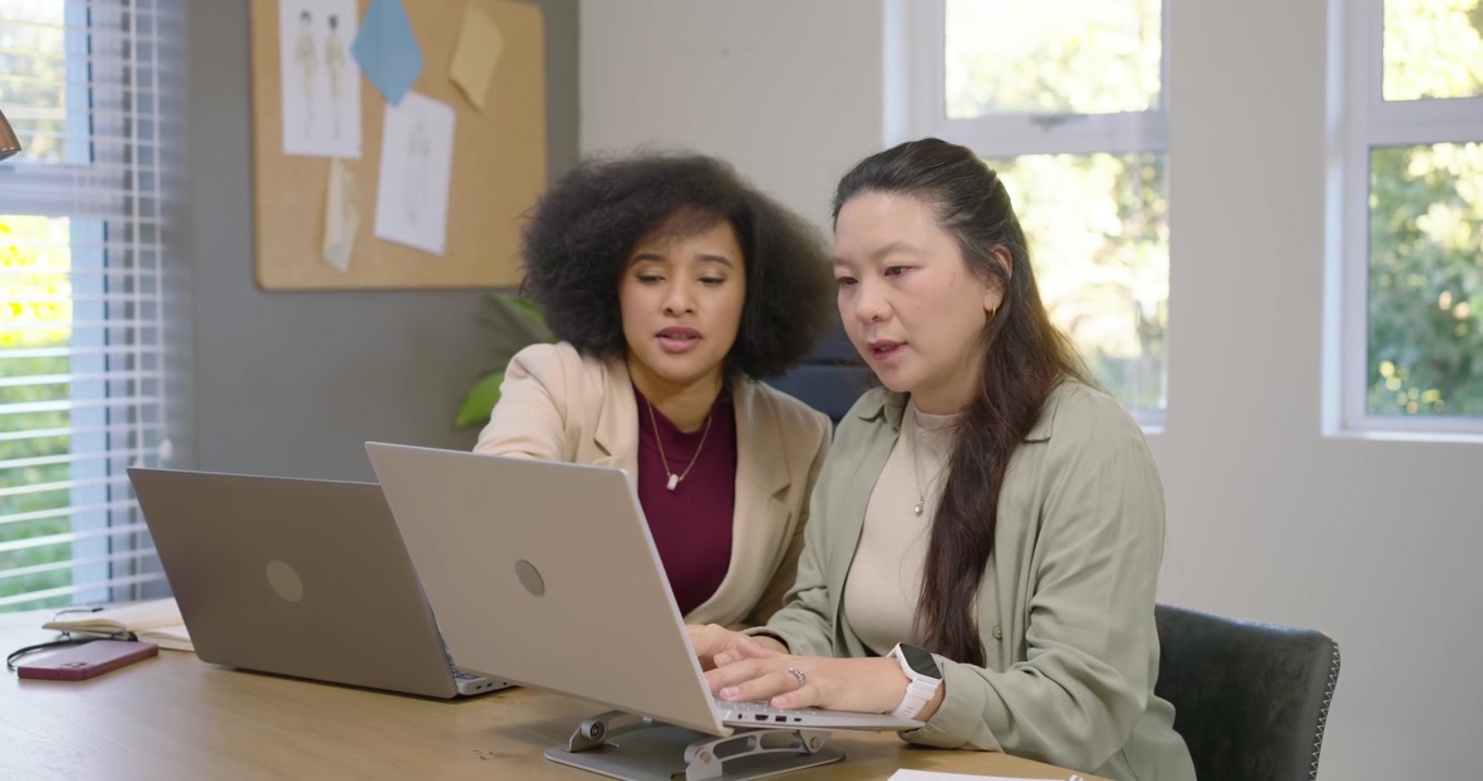 Diverse female duo collaborating at office, right woman pausing, left pointing at laptop on stand