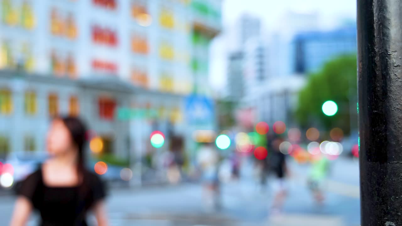 Out-of-focus city street with colorful building, moving pedestrians, and traffic lights, shot during daylight. Camera remains stationary, capturing soft, dreamy urban atmosphere