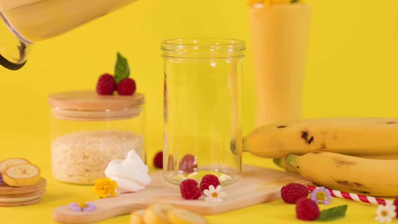 Creamy banana smoothie poured into glass amid tropical ingredients, bright yellow background, static close-up shot