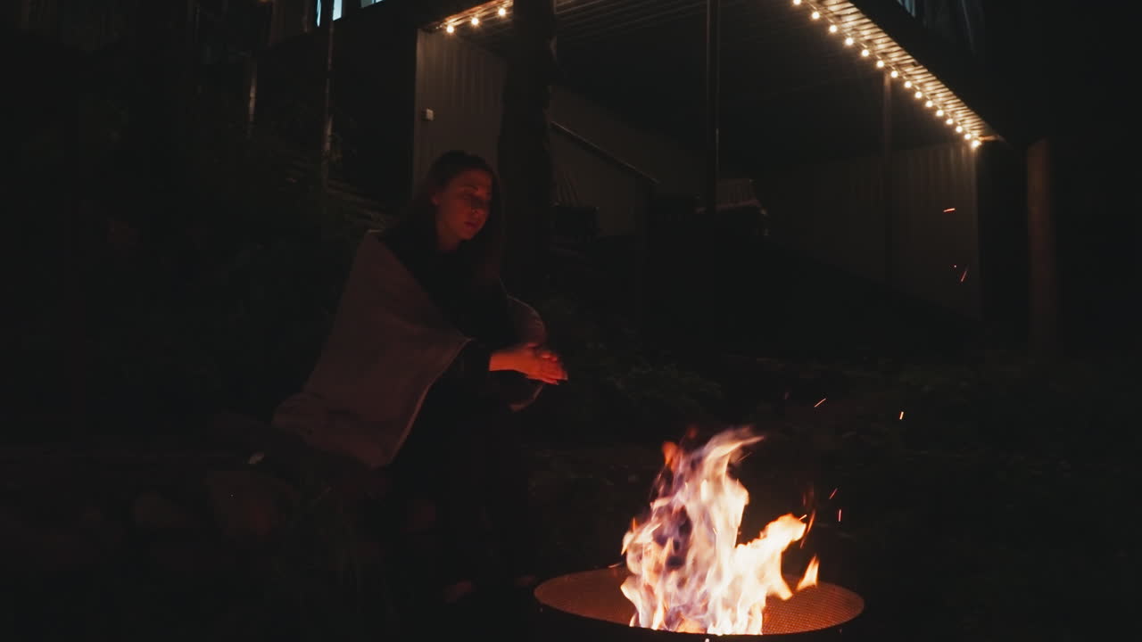 женщина сидит на декоративных камнях у костра. молодая жена с одеялом на плечах держит руки за кострами, сидя в заднем дворе кемпинга вечером