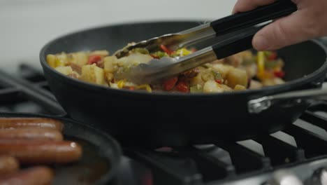 eine hand, die ein paar zangen hält, rührt leicht eine haschmischung aus wurst, pfeffer und kartoffeln in einer bratpfanne