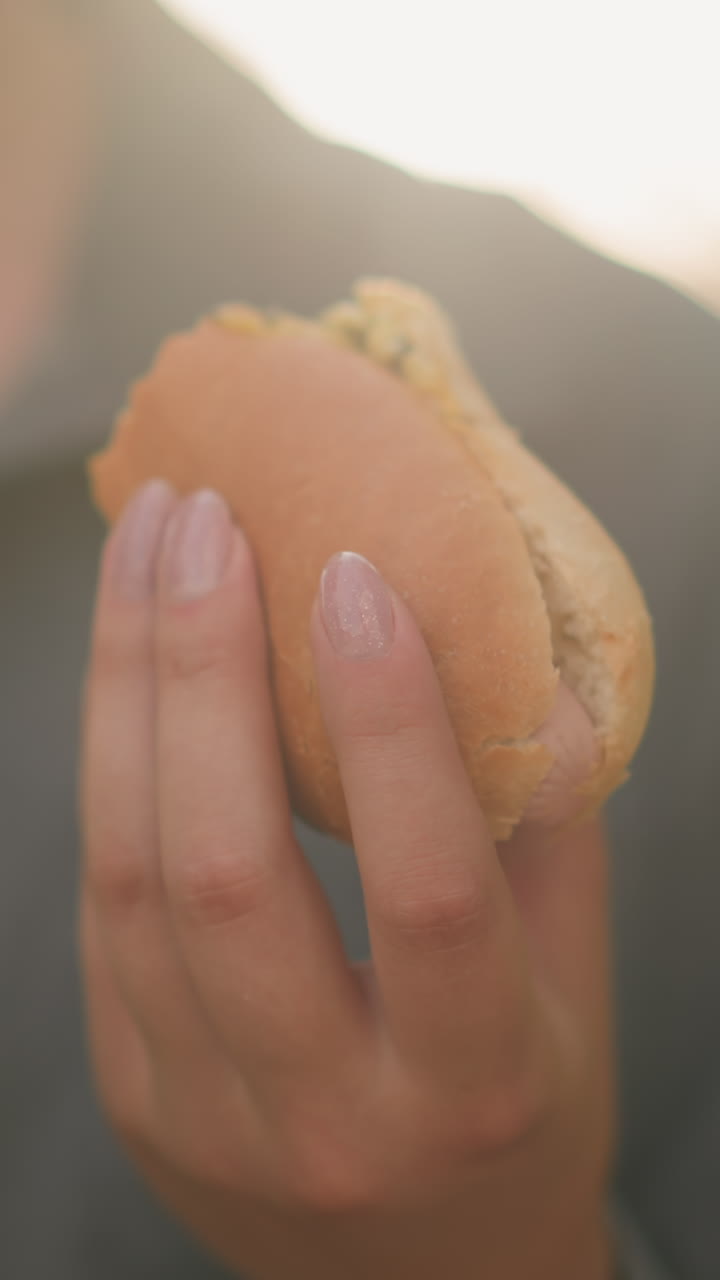 primer plano de una mujer con una camiseta gris sosteniendo un shawarma mientras lee un libro en un entorno al aire libre iluminado por el sol, la luz del sol suave ilumina su mano y el bocadillo, con un fondo borroso de vegetación y cálidos rayos de sol