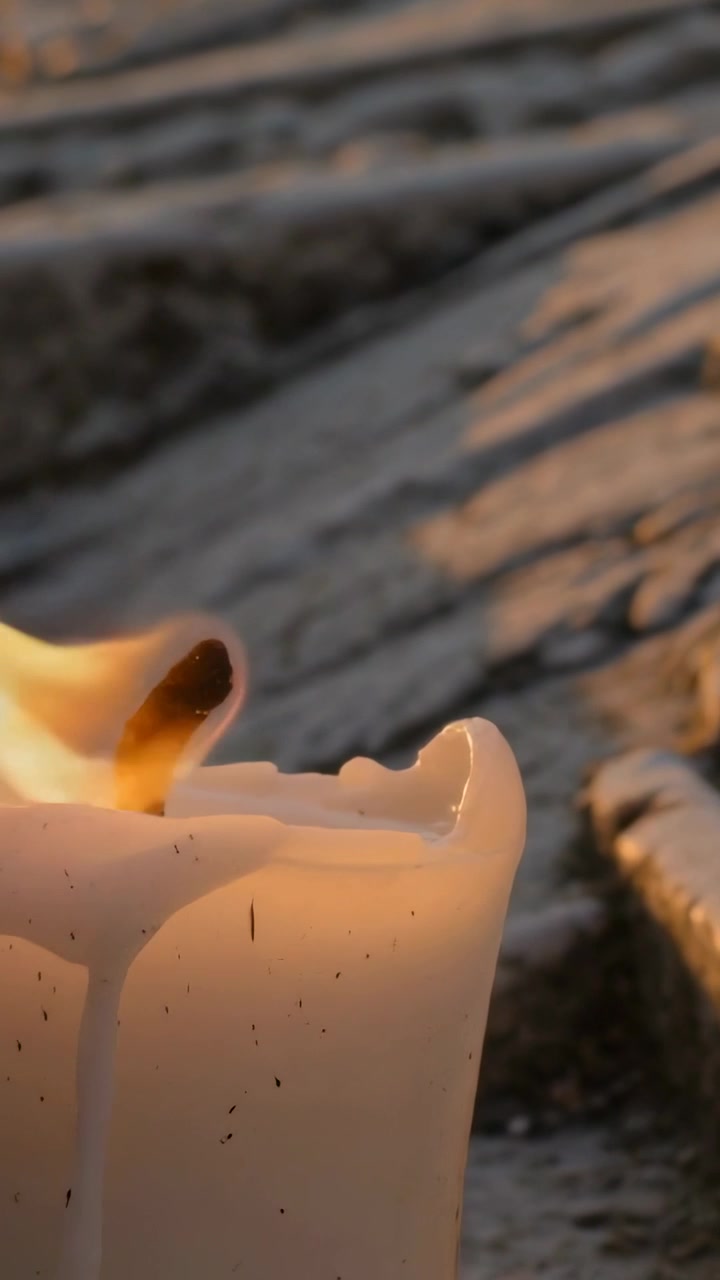 Vertical video: Leaning white candle in wind on stone, wick burning and melting wax, copy space