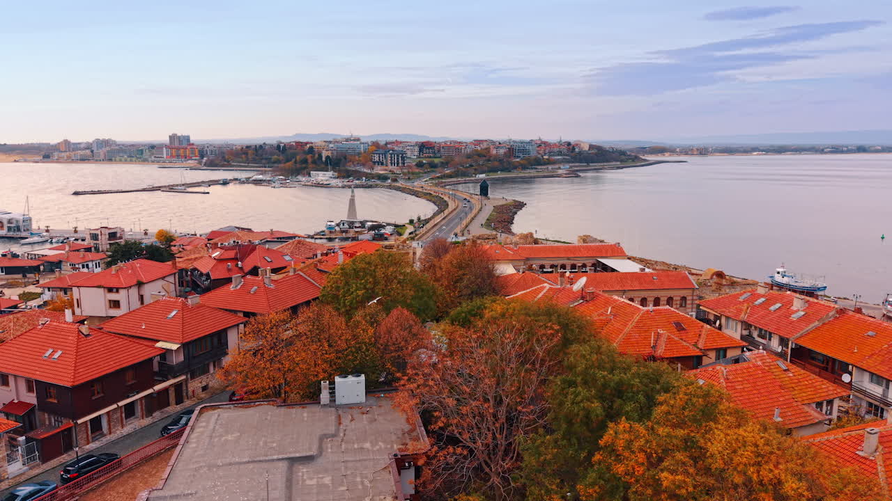 Una città costiera mostra file di edifici con tetti rossi sotto un cielo limpido. La scena cattura la costa con le barche in acqua e il profilo distante di altre strutture.