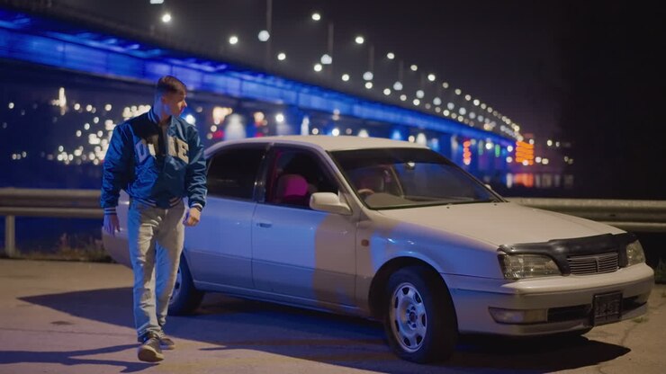 Hombre vestido de forma informal bajo las luces brillantes de un puente con efectos neón reflectantes. Hombre seguro de sí mismo posando junto a un puente iluminado que exhibe reflejos neón cinematográficos en un tranquilo entorno al borde de la carretera.