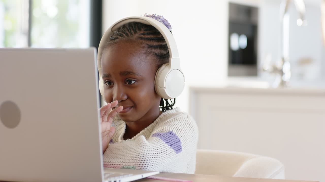 African American girl adjusting headphones, typing on laptop and waving to greet teacher in kitchen