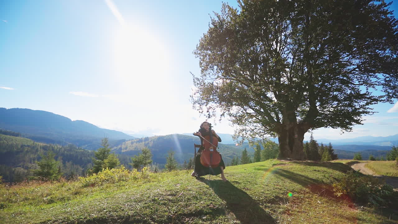 Emotional cello player performing music in the nature. Female musician plays bass-viol sitting at mountainous backdrop.