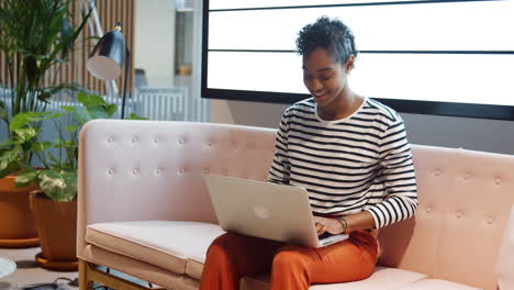 Close-up-of-mixed-race-businesswoman-working-on-a-laptop-computer-sitting-on-a-sofa-in-a-casual-office-smiling