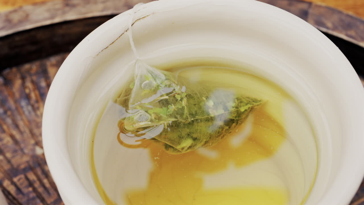 Close up of a woman in a red suit dipping a green tea bag in hot water in a white cup
