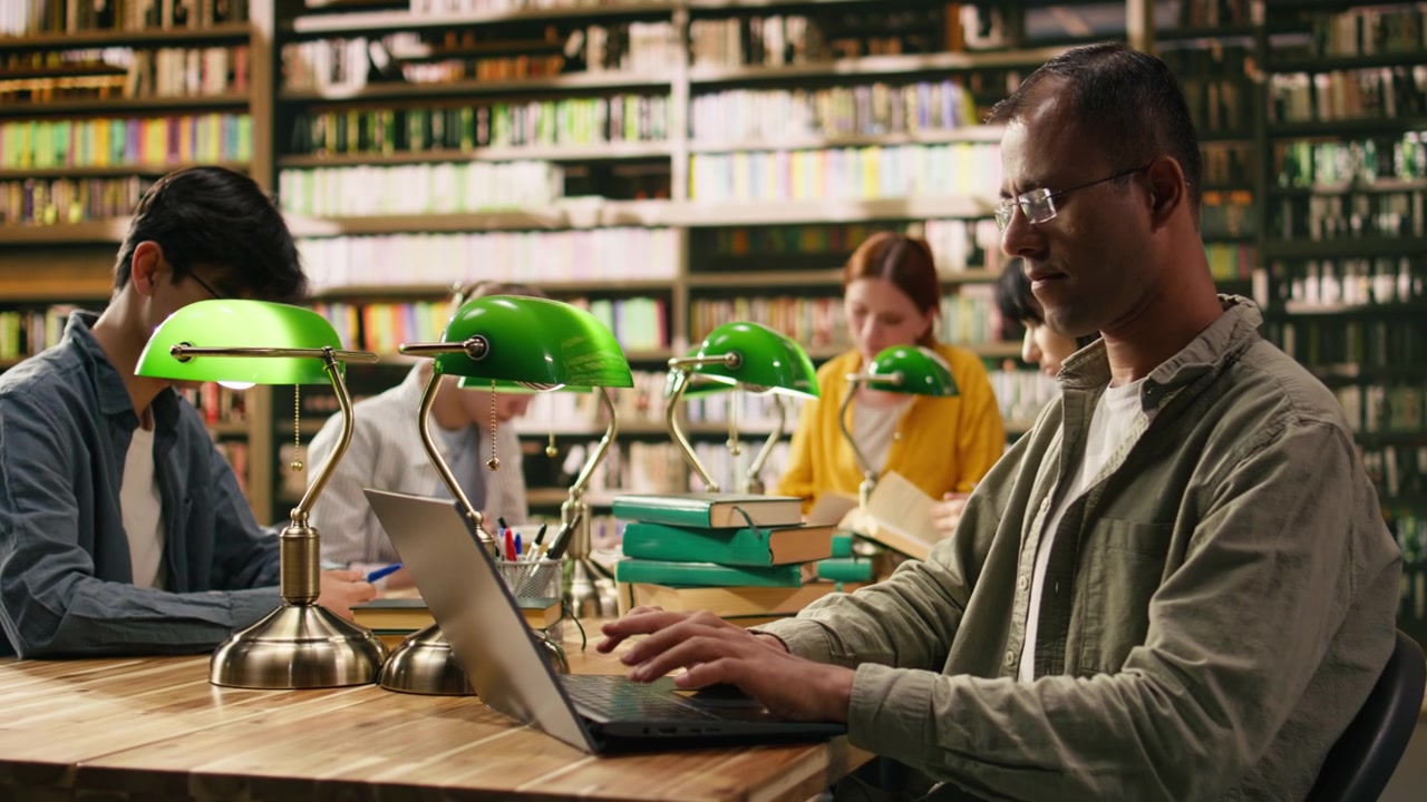 Adult student reading book at a wooden library table with green lamps