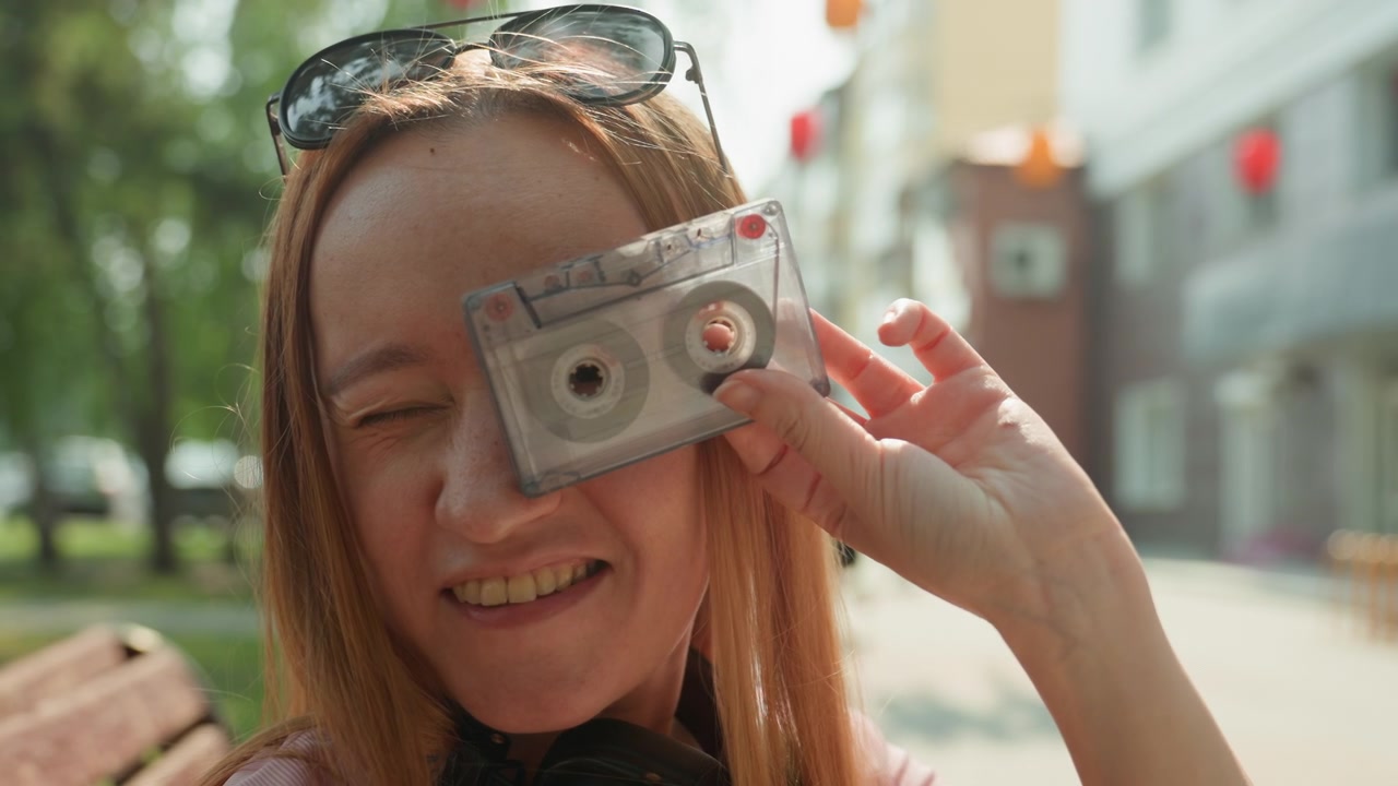 Woman with long red hair holds smartphone and cassette tape in outdoor urban setting wearing sunglasses on head and headphones around neck. Background features green trees and buildings