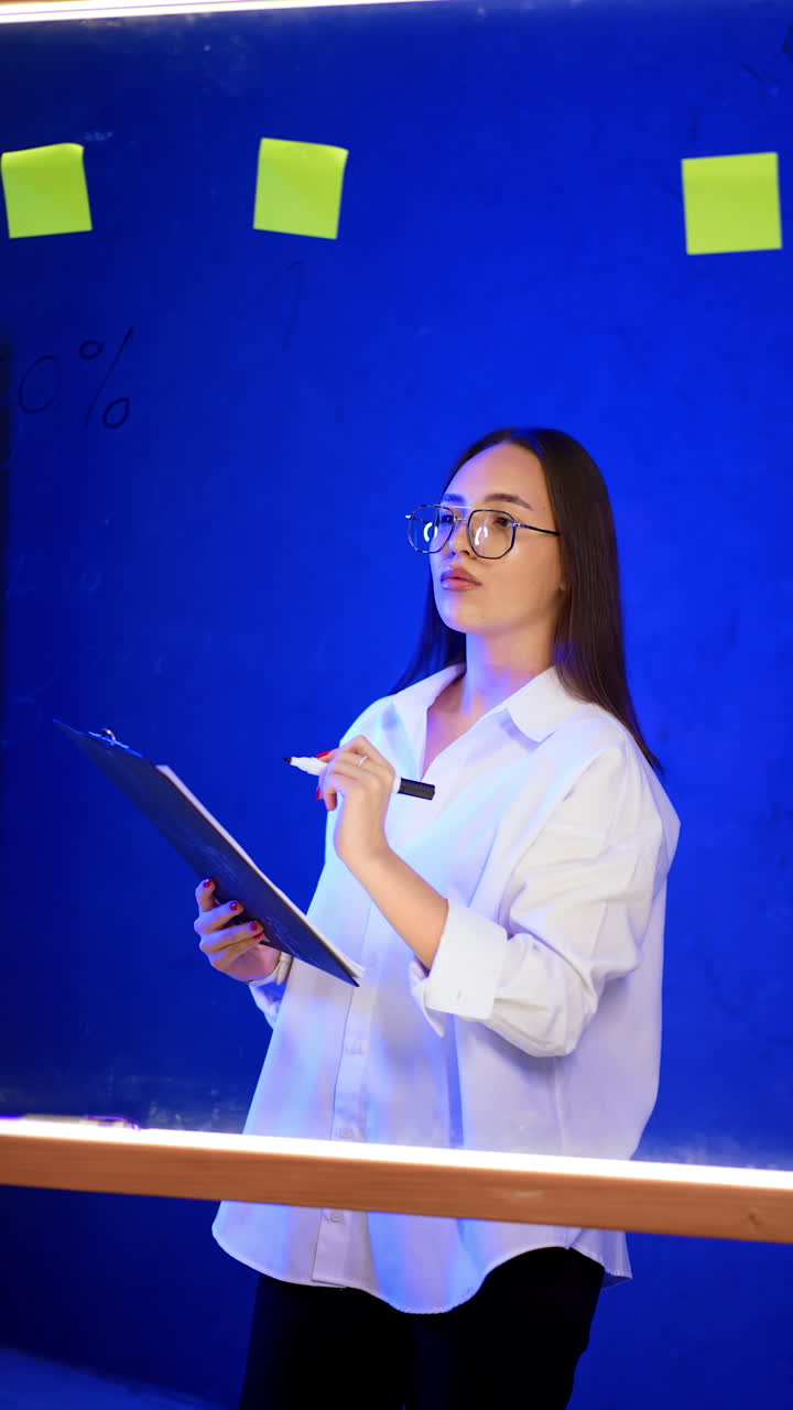 Focused Caucasian brunette lady wearing glasses holds the clipboard. Lady writes on the board with a marker. Vertical video.