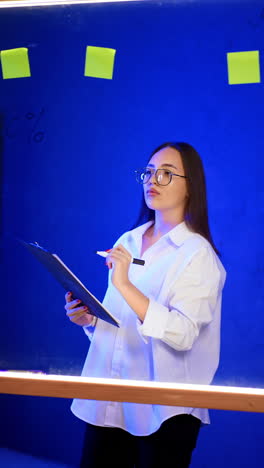 focused caucasian brunette lady wearing glasses holds the clipboard. lady writes on the board with a marker. vertical video.