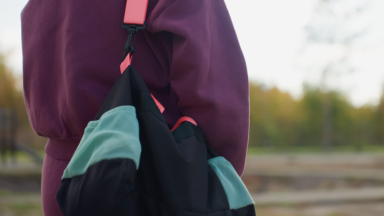 Back view workout participant carrying sport bag over shoulder walking towards modern building along gray tiled pathway beside green lawn under bright daylight with autumn foliage