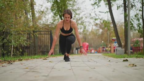 a lady in black fitness wear squats down on a walkway, placing both hands on the ground, prepared to start running, the background includes trees and a park setting