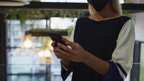 video de una mujer de negocios caucásica con máscara facial usando un teléfono inteligente en las escaleras de la oficina